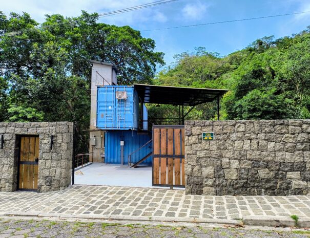 Fachada da casa container dentro do terreno, com acesso pelo portão e área de apoio na entrada.